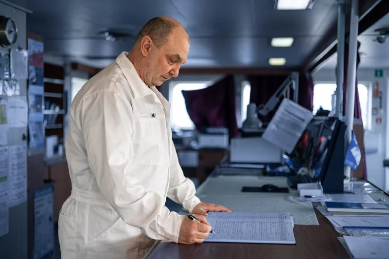 A bald male crew member in a white uniform writes in a logbook on a wooden desk inside a ship's interior.