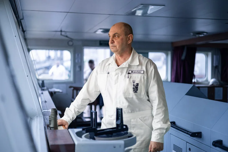 A male ship master in a white uniform stands on the bridge of a vessel, looking out of the window with binoculars on a contro