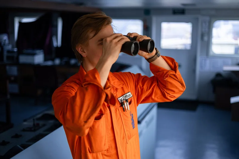 A ship's officer in an orange uniform and '3rd Officer' patch looks through binoculars inside a vessel's bridge.