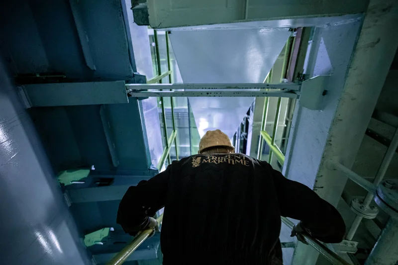 A worker wearing a hard hat and jacket descends a metal staircase with a yellow handrail inside an industrial structure.