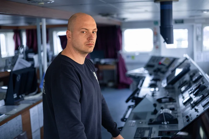 A bald man in a dark blue shirt stands on a ship's bridge, looking at the camera, with a navigation control console in the ba