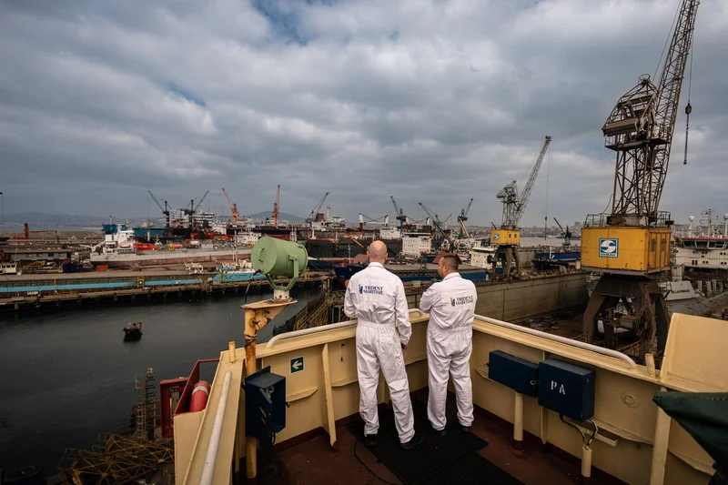 Two men in white jumpsuits stand on a ship deck overlooking a busy shipyard with many vessels, cranes, and dry docks under a