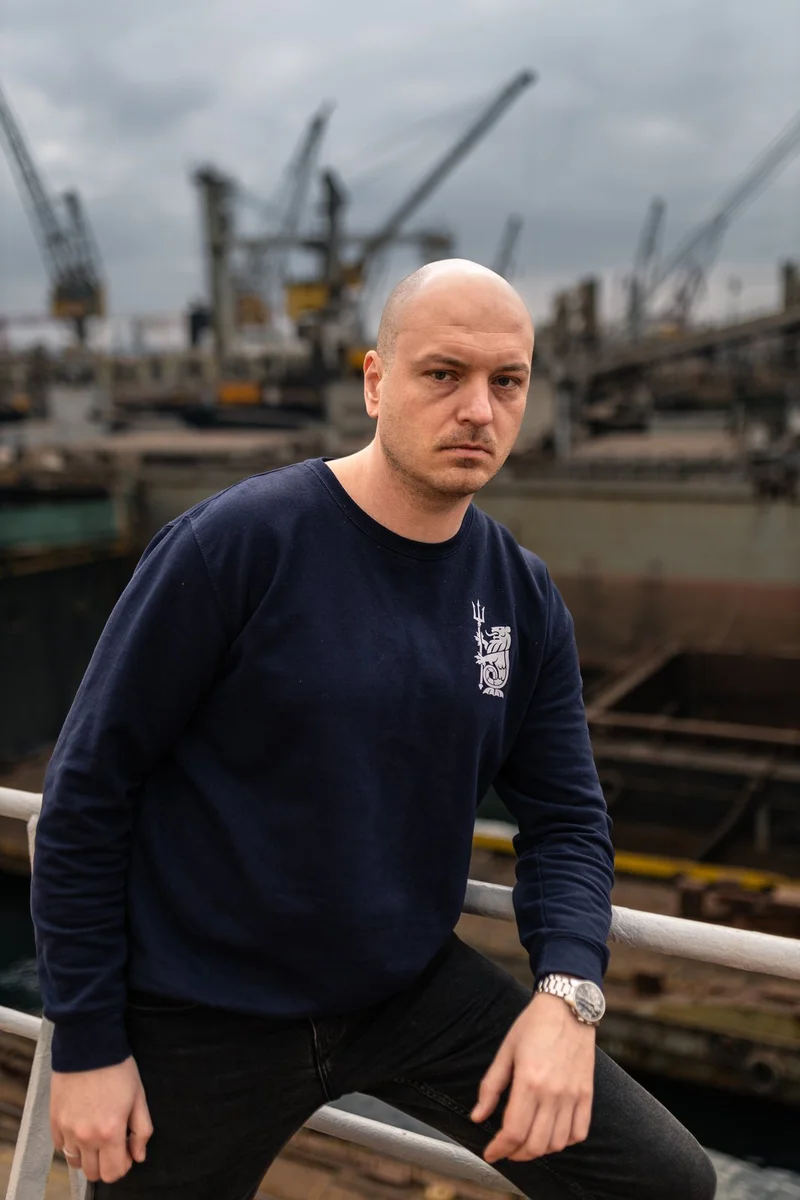 A bald man in a dark sweatshirt leans on a railing with multiple port cranes and vessels visible in a blurry dockyard backgro