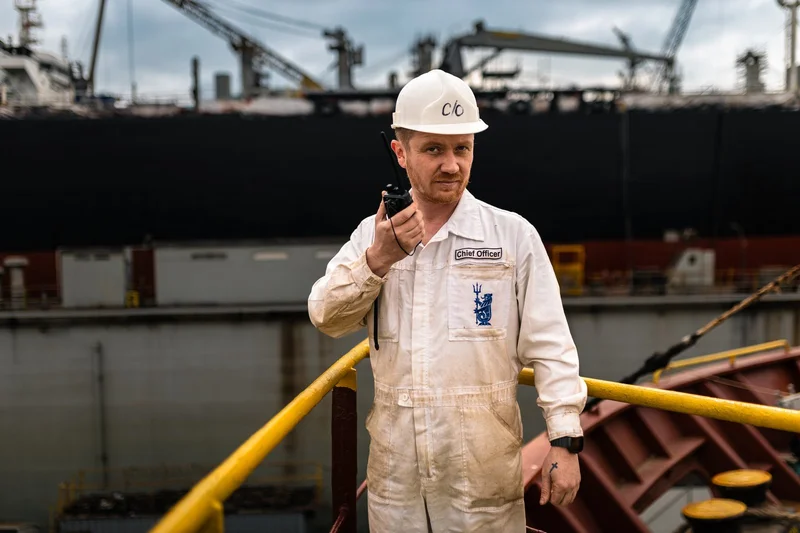 A male Chief Officer in a white overall and hard hat holds a walkie-talkie on a vessel, with a large ship and port cranes in