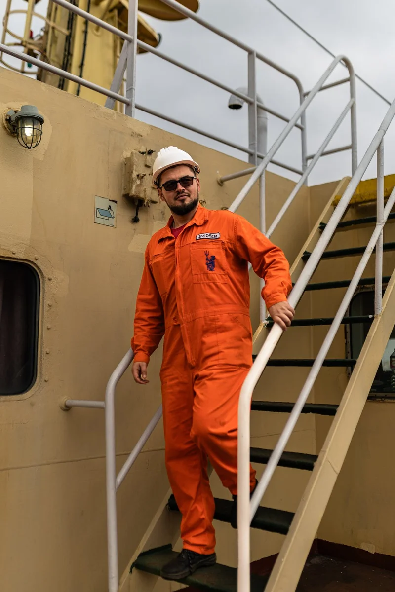 A male 2nd Officer in an orange jumpsuit, white hard hat, and sunglasses descends metal stairs on a ship deck.