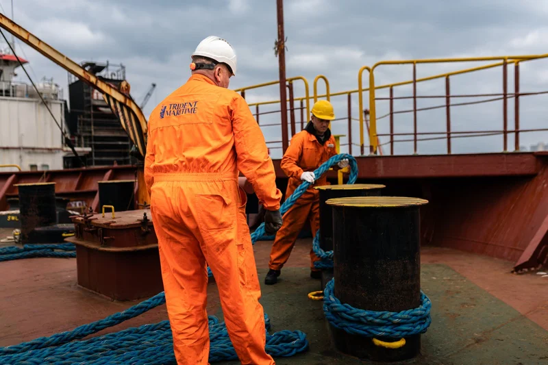 Two crew members in orange coveralls and hard hats handle blue mooring ropes around black bollards on a ship's deck.