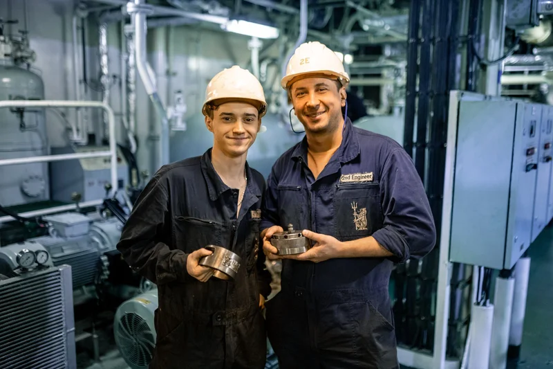 Two male engineers in hard hats and overalls hold mechanical parts inside a ship's machinery room with pipes and equipment.