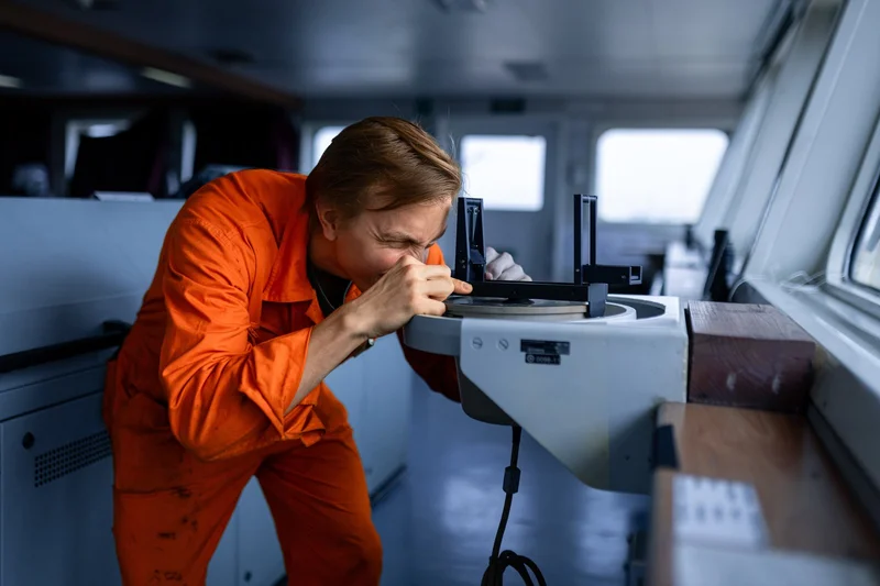 A male crew member in orange overalls uses a navigation device on the bridge of a vessel.