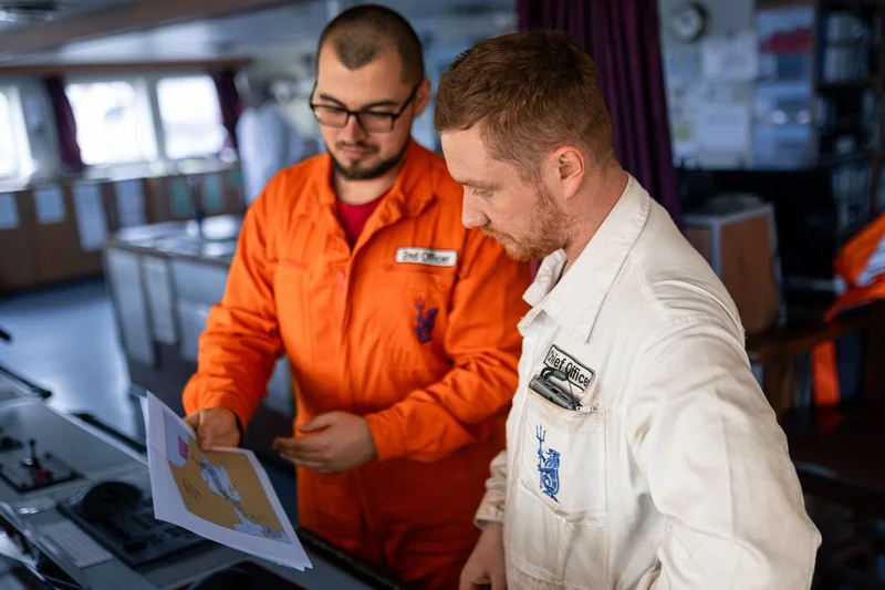Two male crew members, identified as 2nd Officer and Chief Officer, examine a printed map on a vessel's bridge.