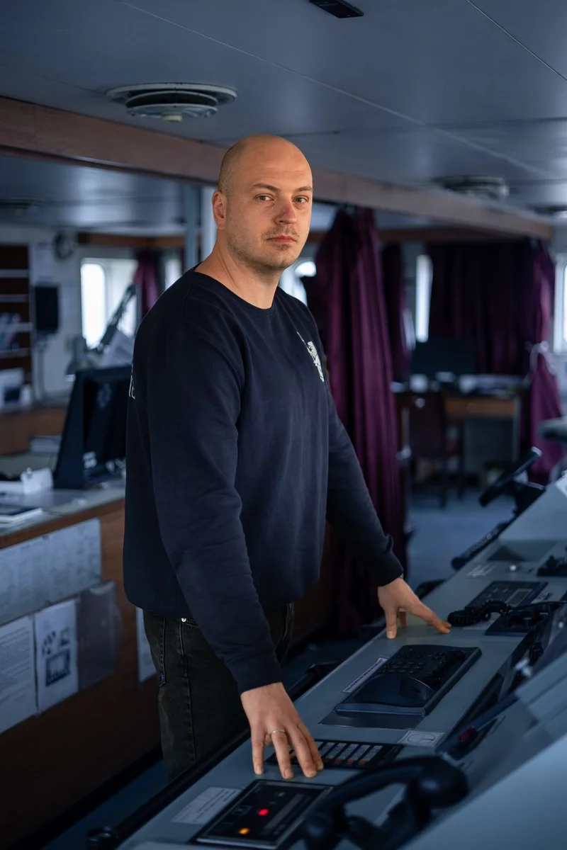 A bald male crew member stands on a ship's bridge, resting his hands on a control panel with buttons and displays.
