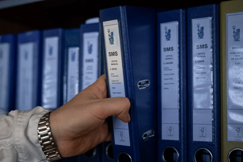 A hand is pulling out a blue "SMS Office Manual 3. DECK" ring binder from a shelf filled with similar manuals.