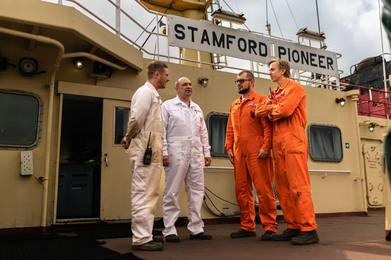 Four crew members in white and orange overalls stand on the deck of the vessel 'STAMFORD PIONEER'.