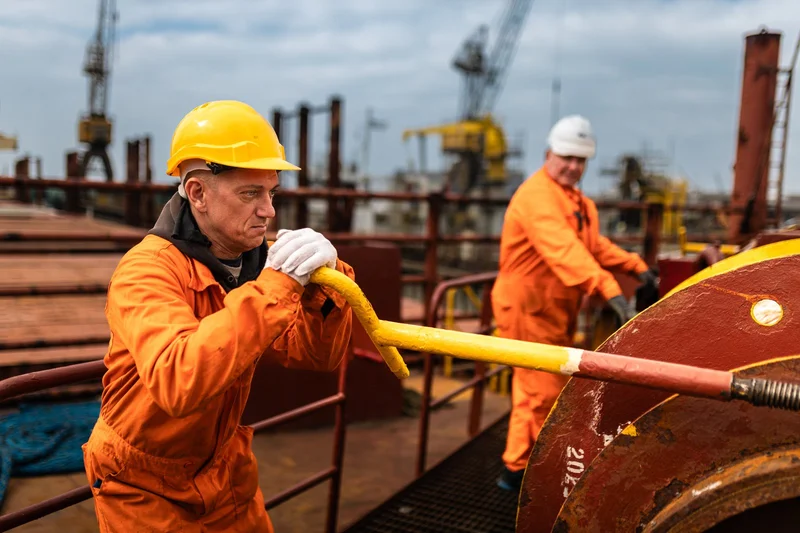 Two crew members in orange coveralls and hard hats operate deck machinery on a vessel, with industrial cranes visible in the