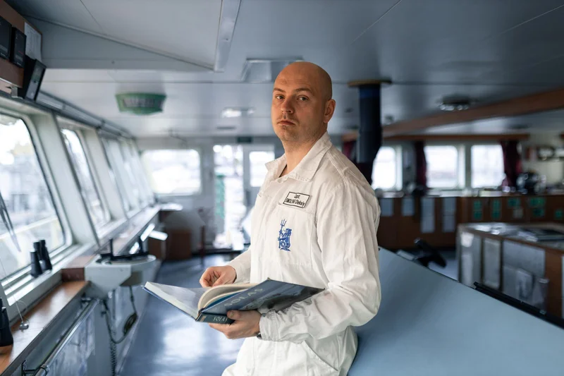 A male crew member in a white uniform shirt holds a SOLAS book on the bridge of a vessel, with windows and navigation equipme