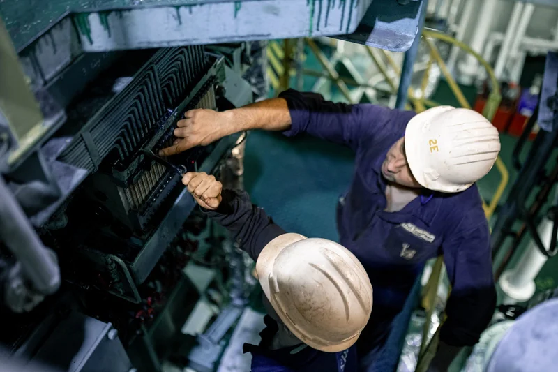 Two crew members in hard hats and overalls are working on a tubular machinery unit with a wrench in an industrial setting.