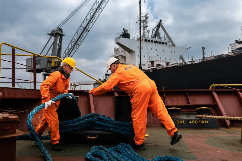 Two crew members in hard hats and orange coveralls handle blue mooring ropes on a ship deck, with port cranes and another ves