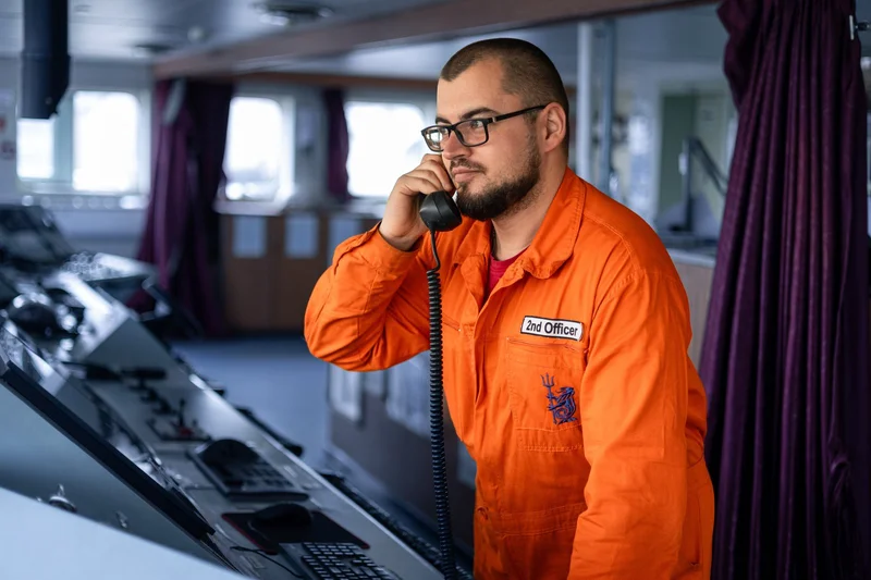 A bearded 2nd Officer in an orange jumpsuit talks on a telephone in a ship's control room with multiple displays.