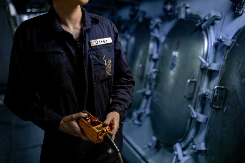 A person in dark overalls holds a yellow control pendant with a red button, standing inside a facility with multiple large me