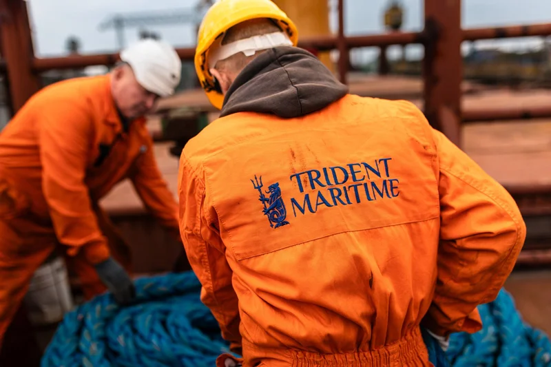 Two crew members in orange coveralls and hard hats are handling a large blue rope on a vessel deck.