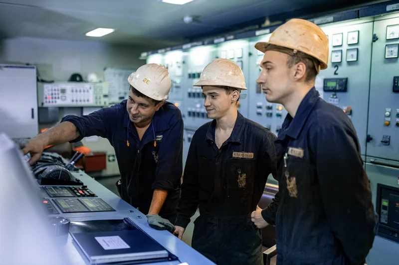 Three marine engineers wearing hard hats and dark overalls are observing and interacting with a vessel's control panel.