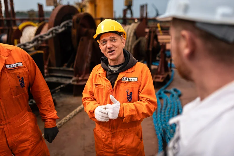 Three crew members, two in orange coveralls and one in a white shirt, are on a vessel deck with winches, chains, and rope vis
