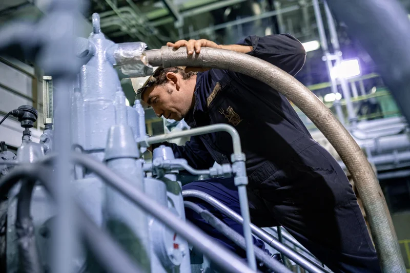 A crew member in a dark uniform and hard hat inspects light blue industrial machinery, holding a large braided hose.