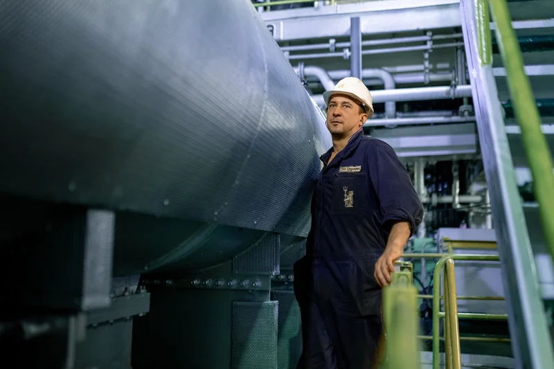 A male 3rd engineer in a hard hat and blue uniform stands next to large machinery and metal stairs in an engine room.