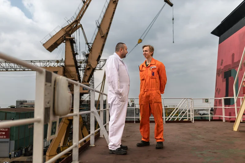 Two men in work overalls stand on a ship deck with a large gantry crane and shipyard facilities in the background.