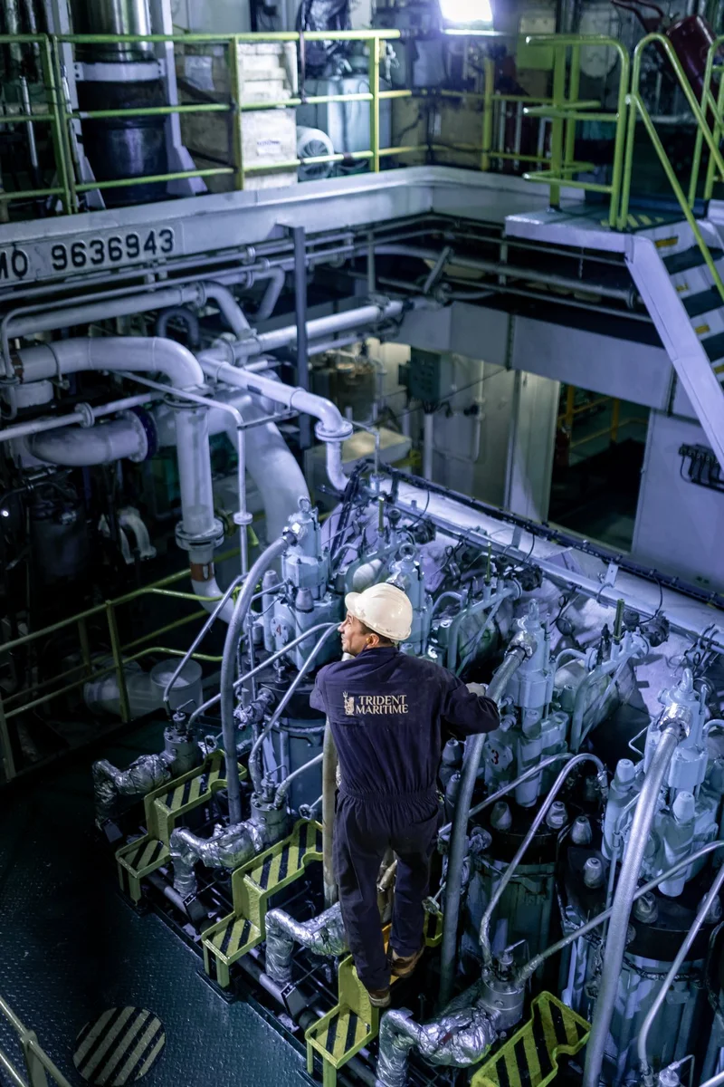 A marine engineer in a hard hat inspects a large vessel engine with pipes and machinery in an engine room.