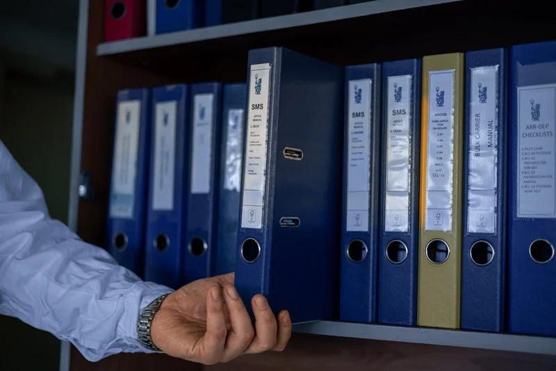 A person's hand reaches for a dark blue binder labeled 'SMS Office Manual' on a shelf filled with maritime operations documen