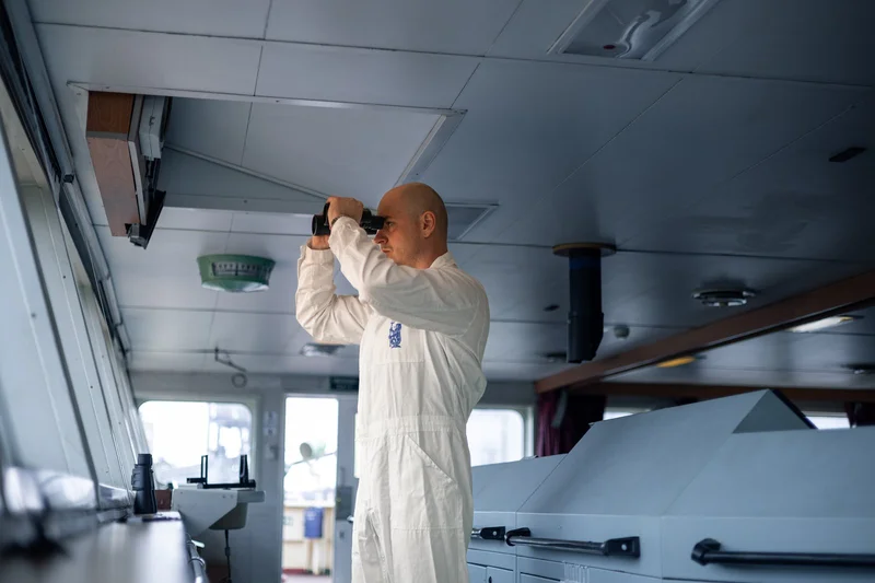 A bald male crew member in a white jumpsuit looks through binoculars on the bridge of a vessel, with control panels and a cei