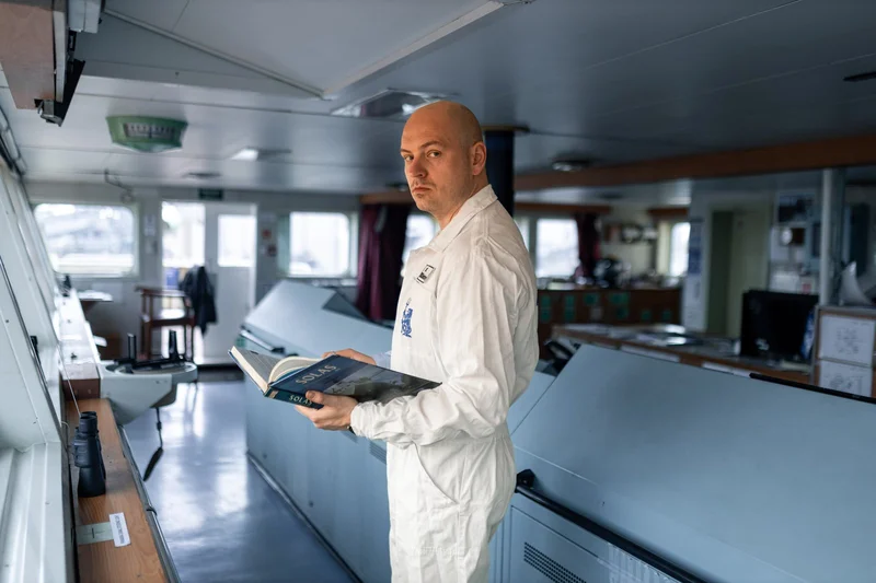 A bald crew member in a white uniform stands on a ship's bridge, holding an open SOLAS book and looking at the camera. Contro