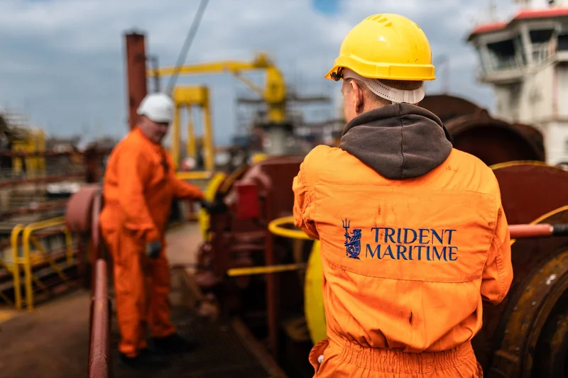 Two workers in orange jumpsuits and hard hats are on a vessel deck, with heavy machinery, industrial cranes, and a ship's bri