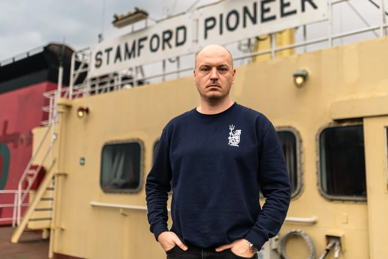 A bald man wearing a dark sweatshirt with an emblem stands in front of the yellow superstructure of the vessel 'STAMFORD PION