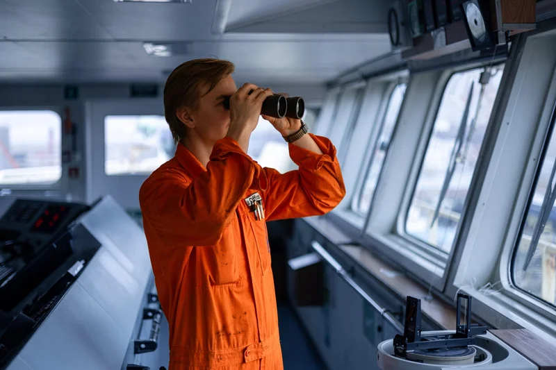 A person in an orange officer's jumpsuit is looking through binoculars on a ship's bridge, with control panels and windows vi