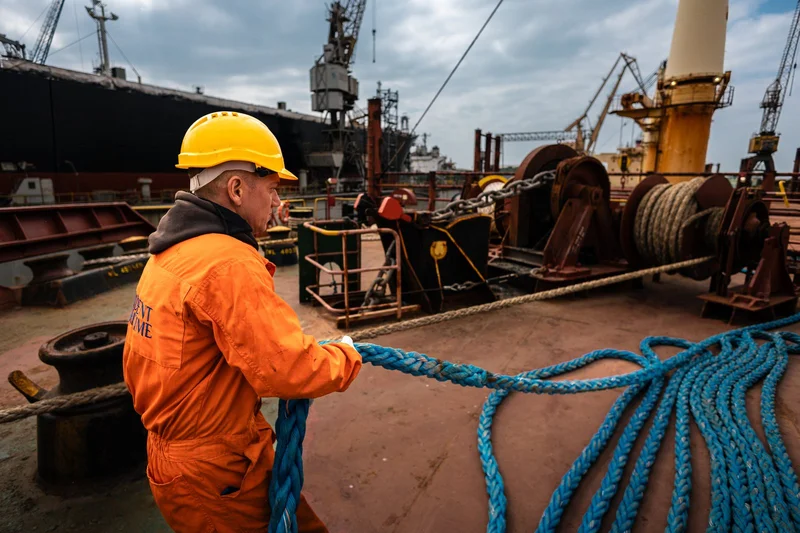A crew member in an orange jumpsuit and yellow hard hat pulls a blue mooring rope on a ship's deck, with winches and port cra