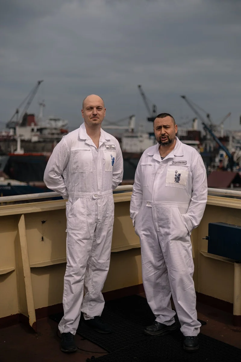 Two men in white overalls stand on a ship deck with other vessels and port cranes visible in the background under a cloudy sk