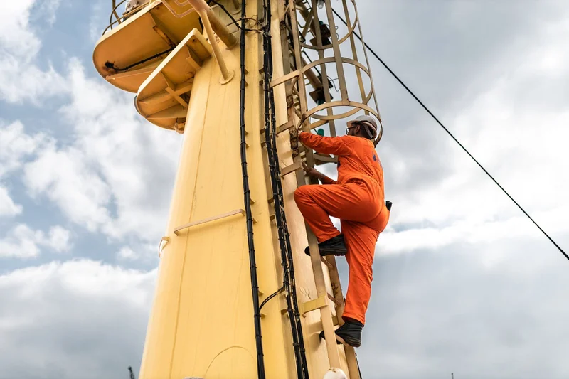 A crew member in an orange jumpsuit and white hard hat climbs a caged ladder attached to a yellow ship mast under a cloudy sk