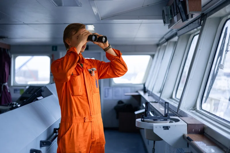 A male crew member in an orange jumpsuit is looking through binoculars on the bridge of a vessel.
