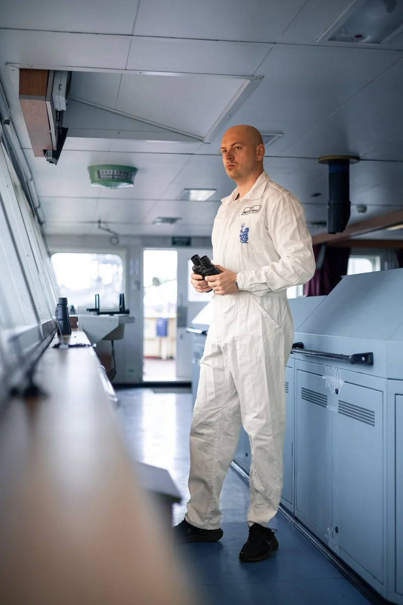 A bald man in a white overall holds binoculars while standing on a ship's bridge with control consoles and large windows visi