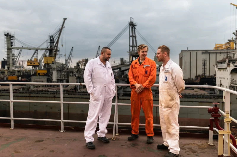 Three men in work overalls are standing on a vessel deck, with a shipyard featuring multiple cranes and industrial buildings